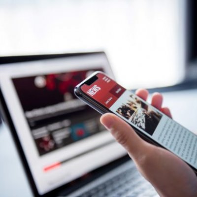 hand holding a phone with screen on a news site in front of  a laptop on a news web page